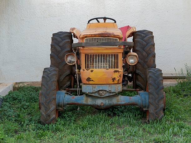 Front view of an old rustic tractor with worn paint and large tires standing on grass near a white plaster wall, symbolizing rural life and vintage machinery.