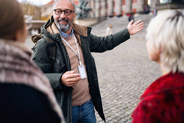 A middle-aged tour guide points with an open hand toward architectural columns while explaining the sights to two young women on a street in Berlin. The scene captures an urban walking tour, cultural exploration, and a casual travel experience in the city.
