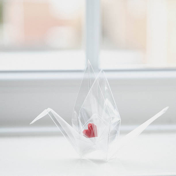 A delicate origami crane folded from transparent acetate plastic featuring a bright red paper heart nestled inside its body, symbolizes fragility, love, and hope through minimalist, modern craftsmanship.
