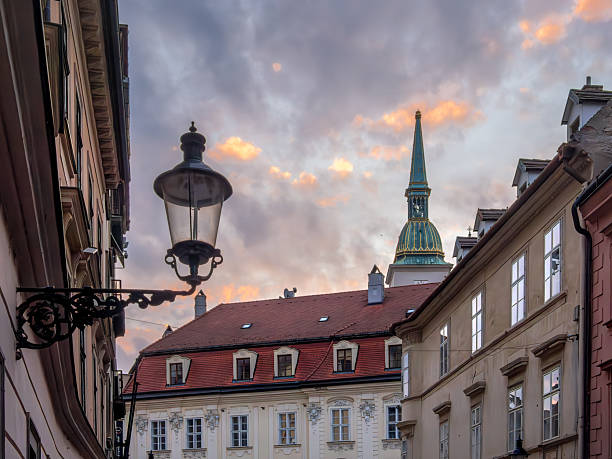 A picturesque street view of Bratislava featuring historical buildings, a lamppost, and a cathedral spire against a colorful sunset sky