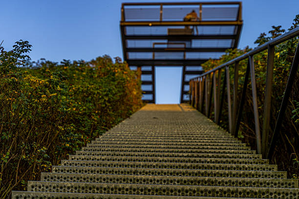 The stairs of perforated metal pull themselves inestopably upwards, their steps merging in an infinite series of targets. On the right, the simple railing accompanies the ascent, while on the left, dense, dark undercog frames the longing for the summit.