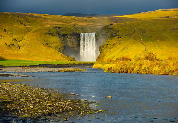 Skogafoss waterfall in Iceland cascades down a steep cliff, creating a powerful mist that forms a vibrant rainbow. The surrounding lush green landscape enhances its majestic beauty