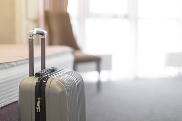 A silver suitcase rests on the floor next to a bed in a hotel room filled with natural light.