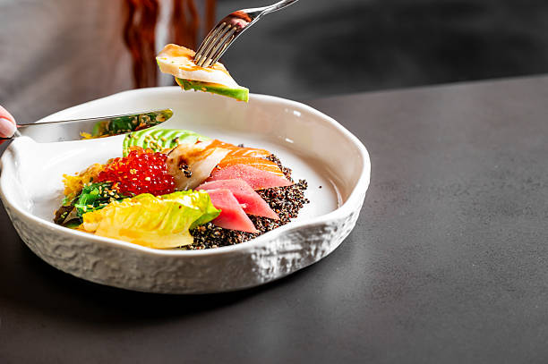 A vibrant poke bowl featuring tuna, salmon roe, avocado, and quinoa, being eaten with a fork and knife on a modern black table