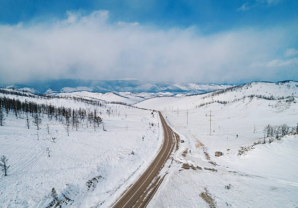 A winding road stretches through a vast, snow-covered landscape, surrounded by gentle hills and dotted with sparse trees.