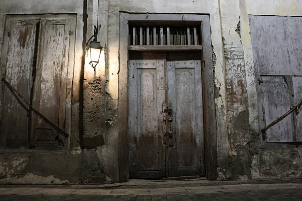 A weathered wooden door in an old building illuminated by a single warm lantern on a rough textured wall showing signs of decay and neglect
