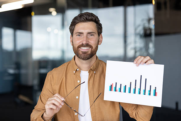istock A man smiles during an online presentation, holding glasses and a chart with graphs, representing data analysis in a modern office environment. 2245499456