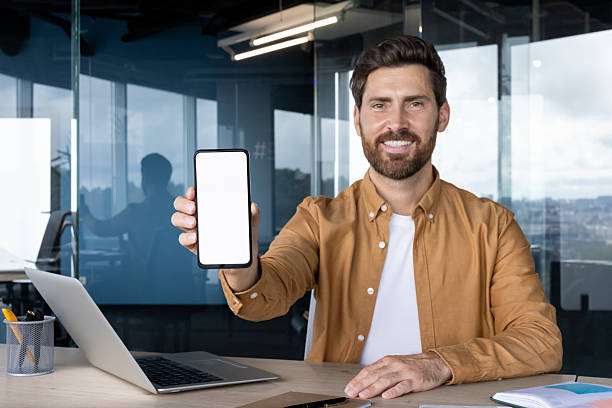 istock A smiling businessman holding a smartphone with a blank screen in an office environment. 2245499728