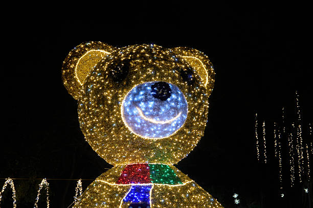 Bright teddy bear face built from LED lights shines against the dark night. Festive holiday decoration with blue nose, golden sparkle, and a cheerful smile.