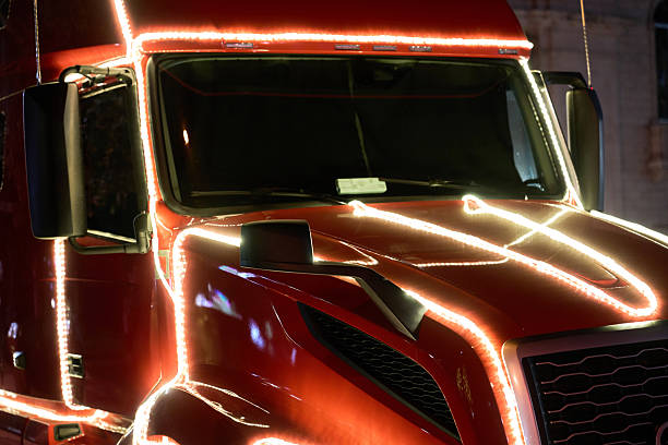 A red tractor trailer cab is traced with glowing white lights along the hood, windshield, and doors at night. The illuminated truck creates a dramatic holiday display on the street.