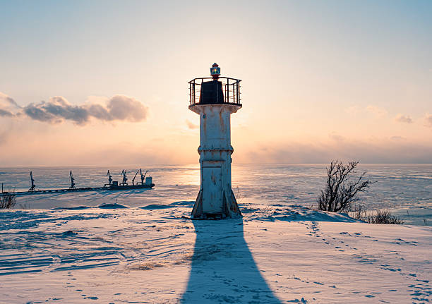 A tall white lighthouse with a metal railing stands on a snowy shore. The sun is setting behind the lighthouse, casting a long shadow across the snow.