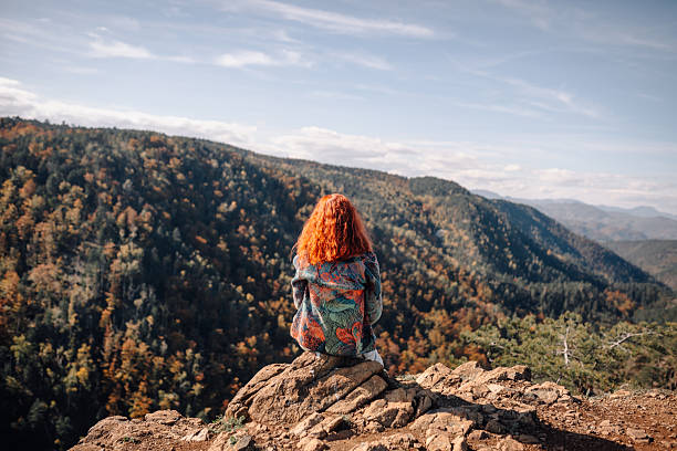 Redhead woman sitting on a rocky cliff enjoying the panoramic autumn mountain landscape and forest, feeling peace and solitude