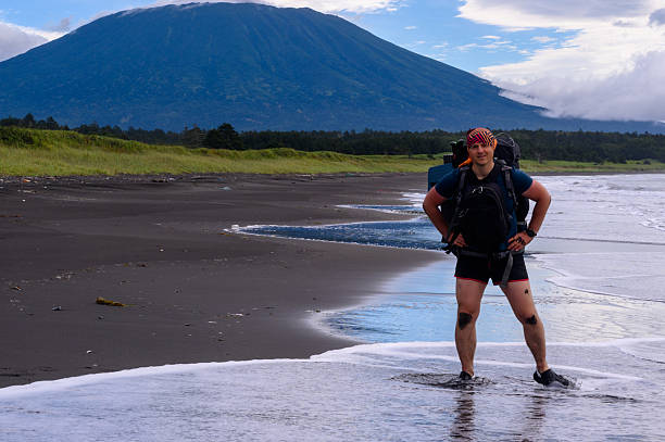 A traveler stands on the wet sand of a beach, surrounded by the beauty of nature. In the background, a majestic mountain rises, and the sky is filled with dramatic clouds.