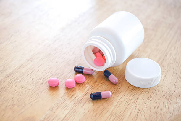 Pink tablets and Capsule spilling from white plastic bottle on wood desk with selective focus.