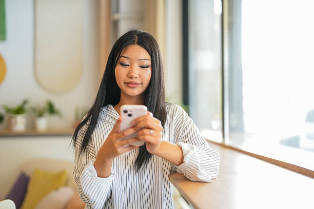Focused young woman holding and using a smartphone while standing by the window in a modern, well-lit home office. Concept of technology, communication, remote work, and digital lifestyle.