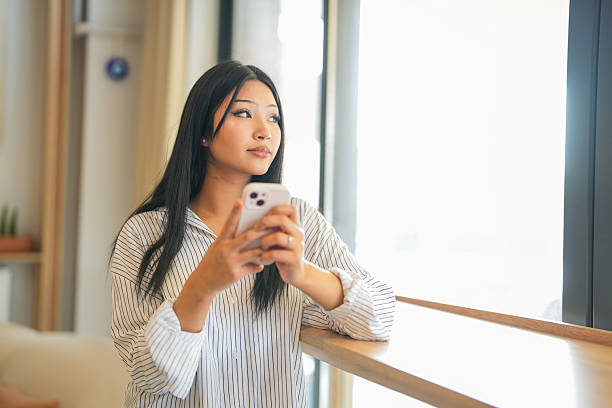 Focused young woman holding and using a smartphone while standing by the window in a modern, well-lit home office. Concept of technology, communication, remote work, and digital lifestyle.