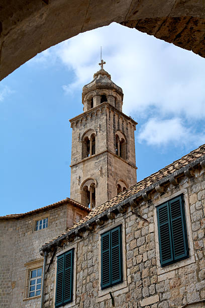 Alleyway perspective in Dubrovnik, Croatia, looking up at a bell tower under a blue sky. Stone buildings with green shutters, a UNESCO heritage site.