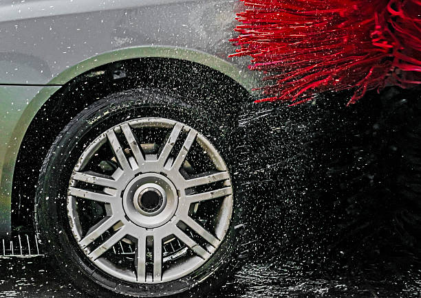 Close-up of silver car wheel being washed in automatic self service car wash with red rotating brushes and strong water spray, detail of cleaning process.