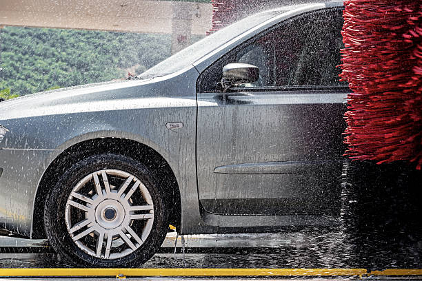 Silver car side view in automatic car wash with red rotating brushes and strong water spray, focus on wheel and door during cleaning process,in a gas station in the Italian Lazio region.