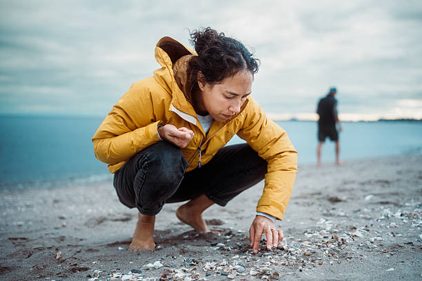 A Eurasian woman wearing a yellow jacket crouches to examine shells and sea glass scattered along the sand at Hammonasset Beach State Park in Connecticut.