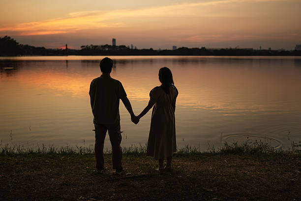A young couple stands in silhouette by a calm lake at sunset, admiring the vibrant sunset reflecting on the water.