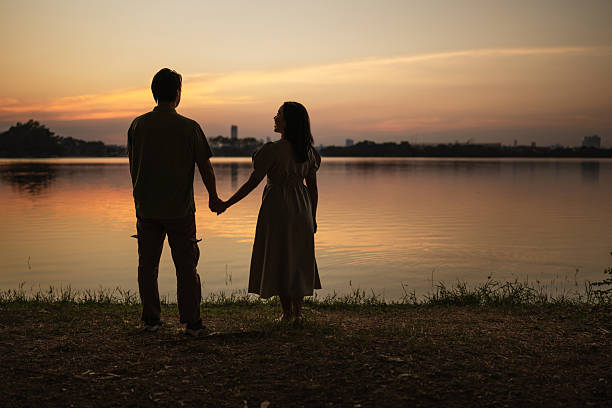 A young couple stands in silhouette by a calm lake at sunset, admiring the vibrant sunset reflecting on the water.