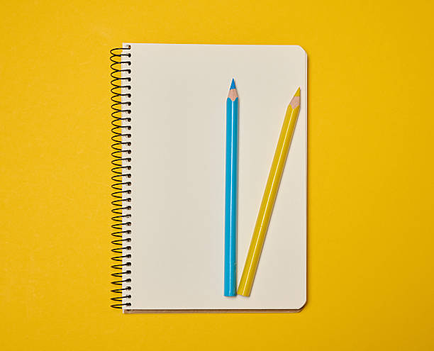istock A spiral-bound notebook with blank pages, topped with blue and yellow pencils, sits against a yellow background. 2245562822