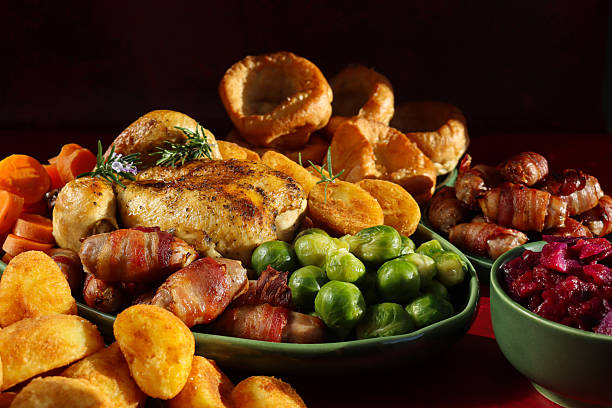 Image of Sunday roast dinner on green serving dish, whole roast chicken with crispy, garnished with Rosemary sprigs, surrounded by side dishes of Brussel sprouts, roast potatoes, sliced carrots, pigs in blankets, braised red cabbage, Yorkshire puddings