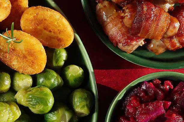 Stock photo showing close-up, elevated view of  a traditional roast dinner served for Sunday lunch on an oval, green serving plate, featuring golden potatoes, pigs in blankets (pork cocktail sausages wrapped in bacon), tender Brussels sprouts, with a bowl of braised red cabbage. This stock image highlights hearty home-cooked meals, comfort food, and family dining.