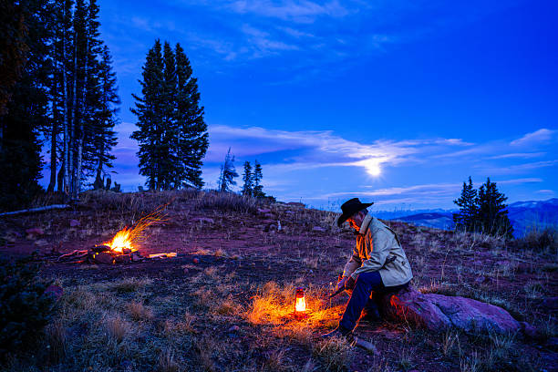 Cowboy man with campfire and glowing oil lantern high on mountain ridge during blue hour dusk. Environmental portrait in autumn with full moon rising in sky in background.
