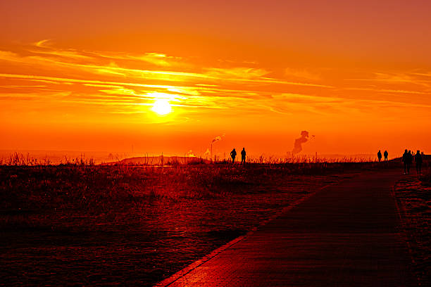 The smooth, red path invites you to face the sunset – or perhaps the morning of a better future. The people on the horizon look like little pilgrims who let themselves be charged by this fiery primaly force of nature. It is the perfect moment when the end of the day becomes a new, shared energy, while the fine smoke flags of civilization quietly rise in the background. A magical place where man and nature meet in the biggest color explosion of the day.
