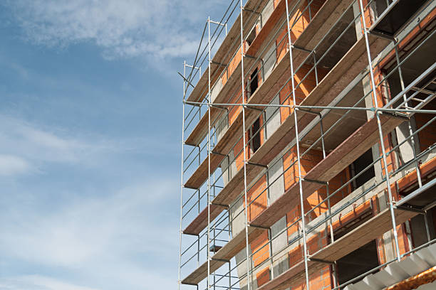 Construction site facade framed by metal scaffolding and exposed orange brickwork, concrete floors and clear blue sky background.