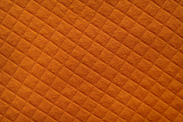 A detailed close-up photograph of orange quilted fabric featuring a diamond stitch pattern, showcasing its soft texture, geometric design, and warm textile surface.