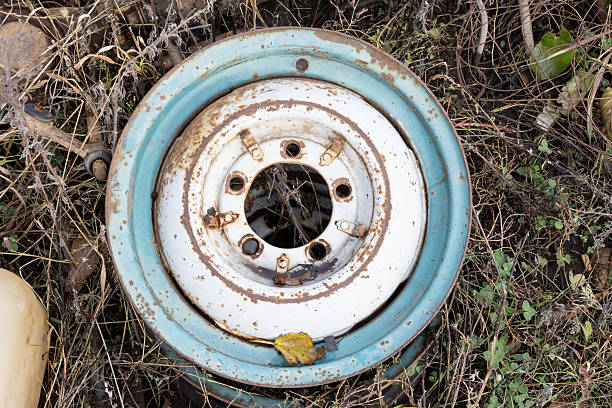 Rusty car wheel rims are piled together at a junkyard. These metal rims, primarily white with significant rust spots, feature multiple circular holes around the center. The area suggests metal recycling and reuse, with grass and scattered debris visible around the stack. Some rims display signs of weathering and decay, indicative of prolonged exposure to the elements.