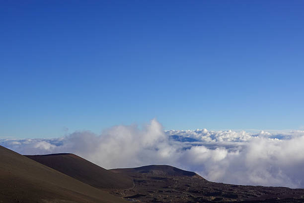 Multiple cinder cones and ridges form dark silhouettes above an expansive sea of clouds, with soft evening light creating gentle gradients of blue and violet. The perspective from high on Mauna Kea emphasizes elevation, remoteness, and the grandeur of Earth’s atmosphere. This image complements adventure, astronomy, or inspirational landscape themes.