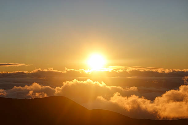 A brilliant golden sun emerges through a gap in the clouds, casting beams of light across dark volcanic ridges at sunset. The cloud tops glow with intense color while the foreground remains in shadow, creating strong contrast and mood. This dynamic sky scene is perfect for motivational themes, adventure travel promotions, or nature-inspired editorial layouts.