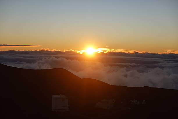 An intense golden sun appears to sink directly into thick swirling clouds, bathing the entire scene in warm light. Dark volcanic slopes frame the bottom of the composition, grounding the otherwise ethereal sky. The photograph captures the dramatic, fleeting beauty of sunset at very high elevations and is ideal for inspirational visuals, travel brochures, and nature themed designs.