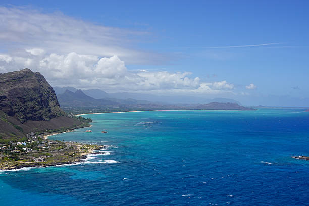 A high-angle view from a hilltop with a Hawaii state flag overlooking the coastal town of Nanakuli, with the blue Pacific Ocean stretching toward the town under a sky filled with fluffy clouds and small patches of bright blue.