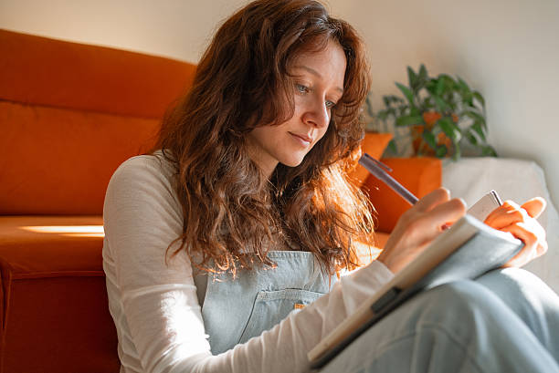 istock Young woman writing notes on notebook while sitting cross-legged on hardwood floor, casual outfit, orange couch blurring background 2245623079