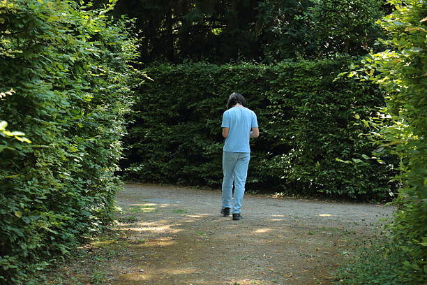 A young man dressed in light blue jeans and t shirt walks along a peaceful garden path surrounded by dense green bushes. The scene captures a calm summer moment in nature with sunlight filtering through the leaves. This image represents solitude reflection and harmony with the environment making it suitable for concepts of mindfulness eco lifestyle and relaxation in nature.