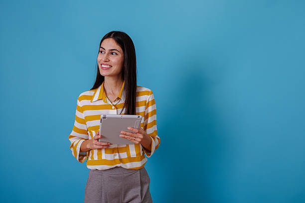 istock Professional woman smiling, holding tablet, thinking about future ideas on blue background with copy space 2245625271
