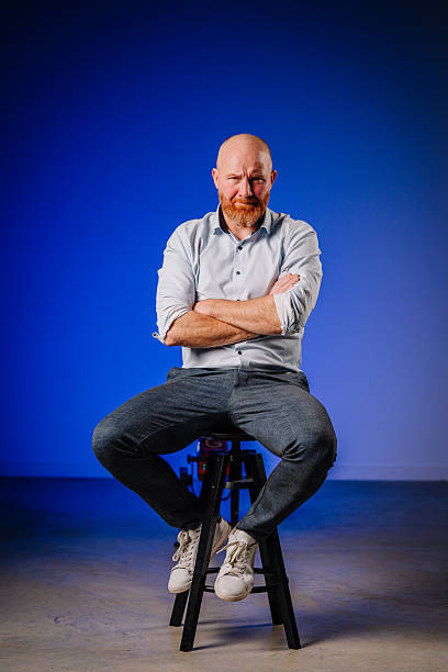 istock Bald man with beard sitting confidently on a stool against a blue background, showcasing a relaxed yet assertive demeanor in a modern studio environment 2245628535