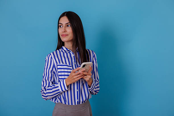 istock Woman holding a mobile phone, looking to the side with a thoughtful expression, standing against a blue background 2245629559