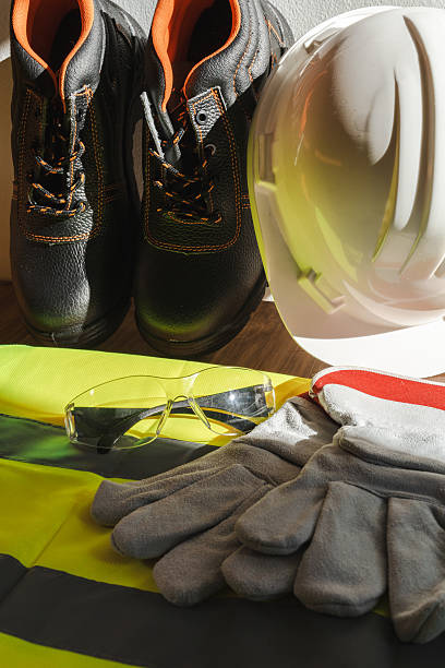 Personal protective equipment (PPE) for safety, showing work boots, hard hat, safety glasses, and gloves on a bright high-visibility vest