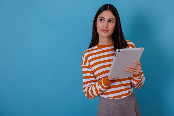istock Young woman using digital tablet, smiling and looking away, standing against blue background 2245635454