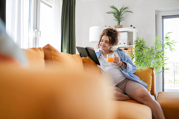 istock Future Mom Sitting Comfortably on Couch with Book Drinking Juice 2245644126