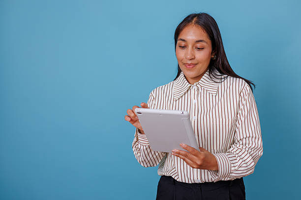 istock Young professional woman happily using digital tablet, browsing communication apps, standing against blue background 2245647388