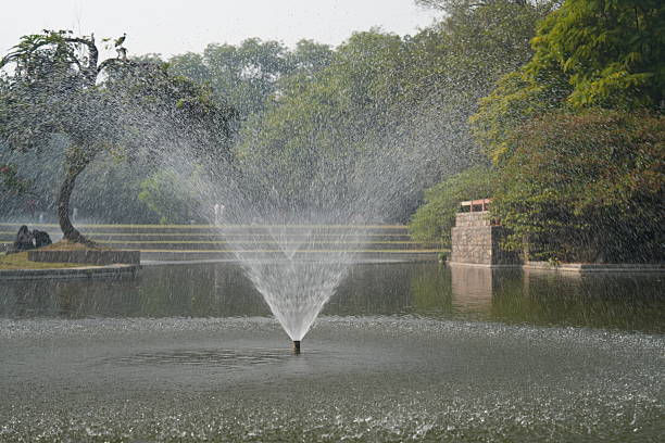 A dynamic burst of water droplets fills the foreground as a fountain operates in a landscaped park. The background trees and shrubs add texture and depth to the composition.