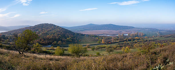 A panoramic view over the rolling hills and wetlands of the Tihany Peninsula, bathed in soft autumn sunlight. The scene captures the diverse landscape of the Balaton Uplands with golden trees, gentle slopes, and a calm blue sky.