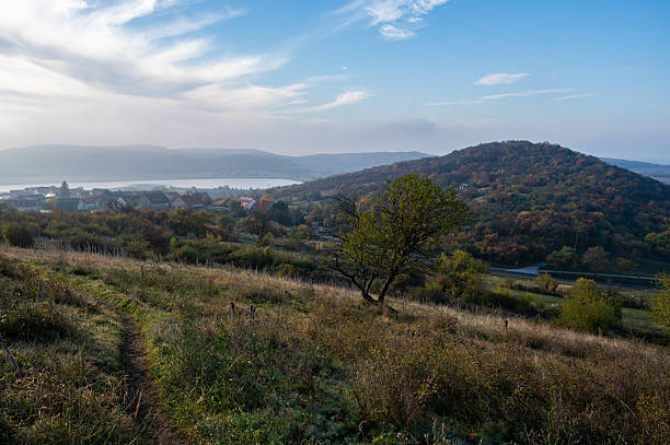 A peaceful autumn morning on the Tihany Peninsula, with soft sunlight illuminating the rolling hills and the village below. The mist over the Lake Balaton creates a dreamy atmosphere, highlighting the natural beauty of this landscape.